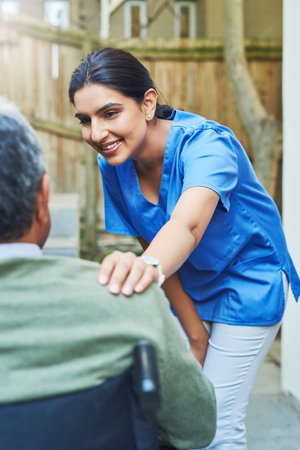 How are you doing today sir. a cheerful young female nurse holding the shoulder of a patient in a wheelchair for support outside at home.の写真素材