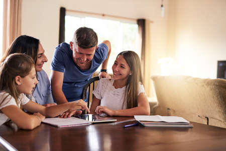 Thanks for the help mom and dad. two cheerful young girls doing homework around a table while getting help from their parents at home during the day.の写真素材