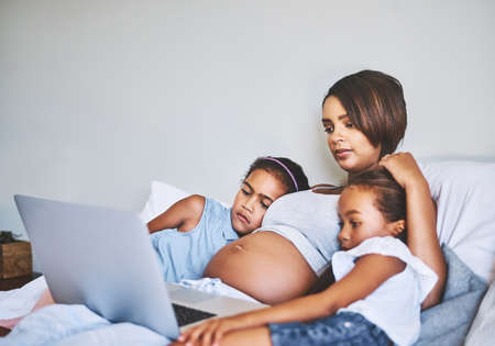 Spending a nice day inside with the kids. two focused little girls relaxing next to their pregnant mother while watching a movie on a laptop at home during the day.の写真素材