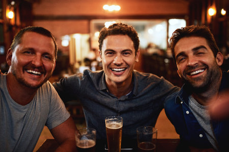 They know each other since childhood. Portrait of a cheerful young group of friends having beers together while being seated at a table inside a bar at night.の写真素材