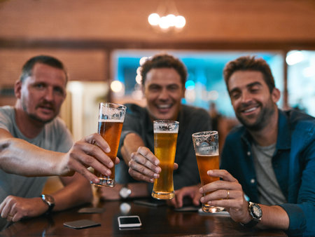 We make the best beer in town. Portrait of three cheerful young men having a celebratory toast with beer while sitting at a table inside of a beer brewery during the day.の写真素材