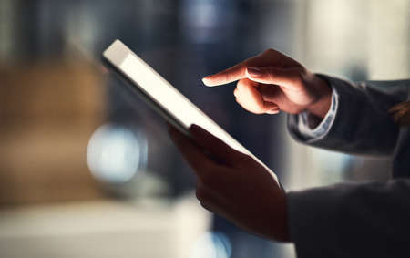 Closeup of woman reading social media news on a digital tablet while working late in an office. Professional female browsing online, researching creative plans and strategies for a business projectの写真素材