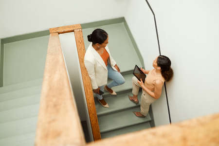 Colleagues discuss, plan and share ideas on a staircase at work in a startup business. Overhead of Businesswomen talking, chatting and socializing on a stairway during a break at a corporate jobの写真素材