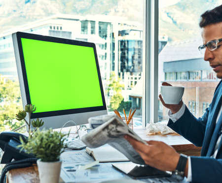 Starting off his day with coffee and the newspaper. a focused young businessman seated at his desk while drinking coffee and reading a newspaper in the office.の写真素材