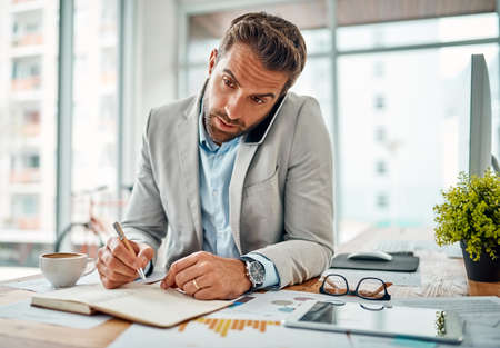 Scheduling all his meetings. a handsome young businessman writing notes while talking on a cellphone in an office.の写真素材