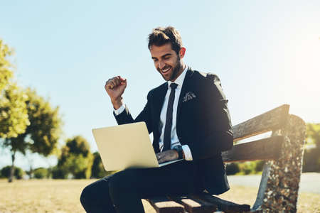 Yes This is fantastic news. a handsome young corporate businessman using a laptop at a public park.の写真素材