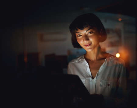 Staying updated on all things business. a young businesswoman using a digital tablet during a late night at work.の写真素材
