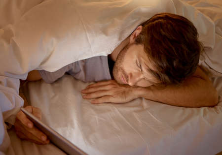Man relaxing in bed on a digital tablet browsing on social media or networking on the internet. Male addicted to technology scrolling on mobile at night in a room. Guy searching the web with a tablet.の写真素材