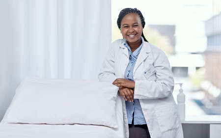 Happy doctor leaning on a hospital bed smiling, standing and ready to serve, assist and help you get healthy. Cheerful, friendly and young medical professional inside an emergency healthcare facilityの写真素材