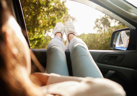 Relaxed, content and happy woman relax while enjoying the sunny morning in a car. Back view of female with feet out her window taking a break from driving sitting and enjoying the summer sunshine dayの写真素材