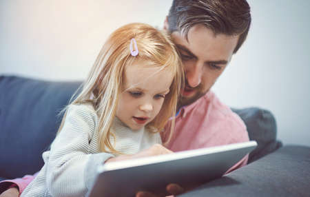 Bonding time in the age of the app. an adorable little girl using a digital tablet with her father on the sofa at home.の写真素材