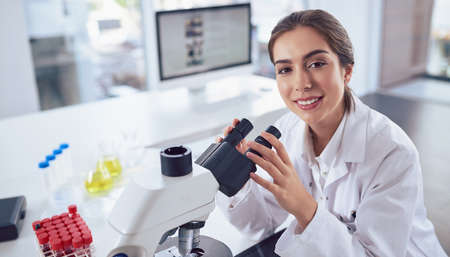 Scientists always have the most state of the art equipment. Portrait of a cheerful young female scientist using a microscope while looking at the camera inside of a laboratory.の写真素材