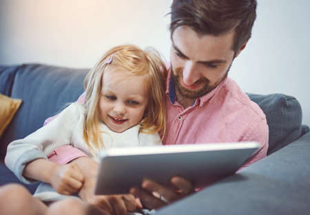 Parenting in the age of digital technology. an adorable little girl using a digital tablet with her father on the sofa at home.の写真素材