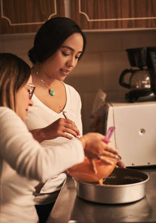 Gently pour mixture into pan. two determined young women baking together in a kitchen at home.の写真素材