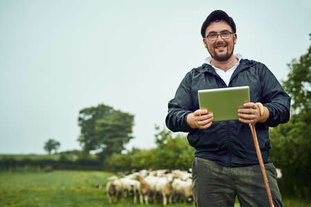Farmers need some technology too. Portrait of a cheerful young farmer standing with a digital tablet and cane while a flock of sheep grazes in the background.の写真素材