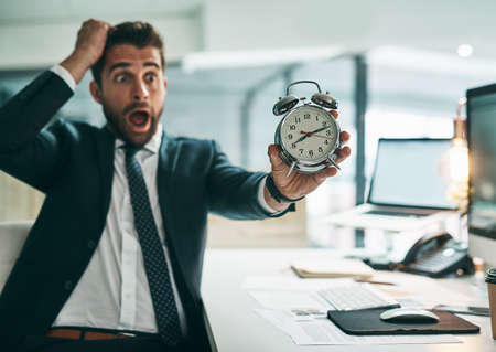 Oh dear is that the time already. a surprised young businessman holding an alarm clock while being...