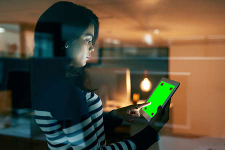 Maintaining her online presence as a business. a young businesswoman working late on a digital tablet with a chroma key screen in an office.の写真素材