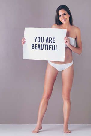 Just sending you a reminder to say...Studio portrait of an attractive young woman holding a sign that reads you are beautiful against a pink background.の写真素材