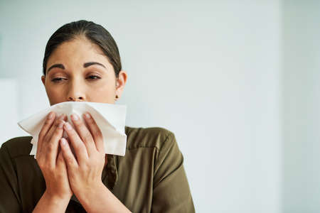 The sneezes just keep on coming. a young businesswoman blowing her nose in an office.の写真素材