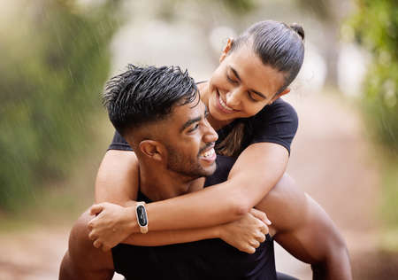 Wet, fitness couple playing in the rain during fun romantic outdoor training, running or healthy workout exercise in park. Happy sport people with a smile or caring man giving woman a piggyback rideの写真素材