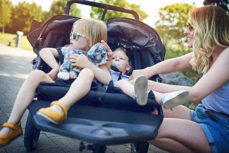 Is everyone all strapped in. a young woman putting her daughters into a pram on a day outdoors.