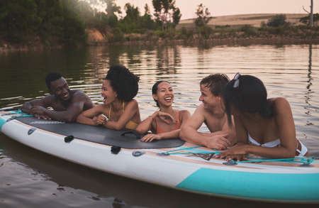 Friends, vacation and having fun while leaning on a paddle board and talking in a lake. Happy and...