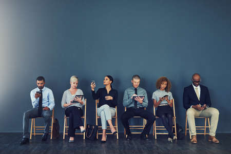 Now comes the wait. Studio shot of a group of businesspeople using wireless devices while waiting in line.の写真素材