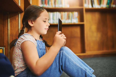 I think its time to get glasses. a focused young girl browsing on a digital tablet while being seated inside of a library during the day.の写真素材