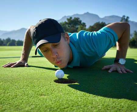 Why wont you go in. Low angle shot of a carefree young man blowing a golf ball into a hole on a golf course.の写真素材