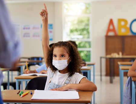 Covid, learning and education with a young girl student wearing a mask and raising her hand to ask or answer a question in class at school. Female child asking or answering the teacher in a classroomの写真素材