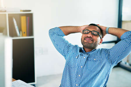 What a day. a handsome young man sitting with his hands behind his head in the office.