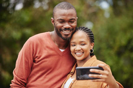 Trendy, modern couple taking selfie on phone while on a romantic date in a park and sharing relationship status on social media app. Smiling, in love married husband and wife capturing happy memoriesの写真素材