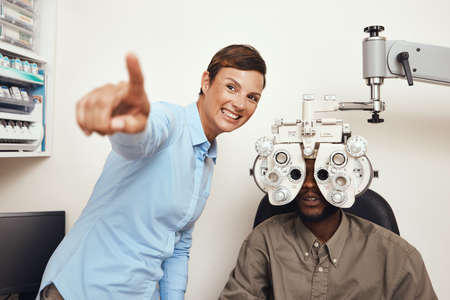 Happy optometrist doing eye test on a patient, examining vision and doing optical exercise at an optometry checkup. Young black man consulting with an opthalmologist, measuring and checking eyesightの写真素材