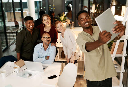 Excited, happy and smiling team of corporate workers taking fun selfie on a tablet in office. Fun, playful and cheerful marketing team posing for social media picture in their new startup agency jobの写真素材