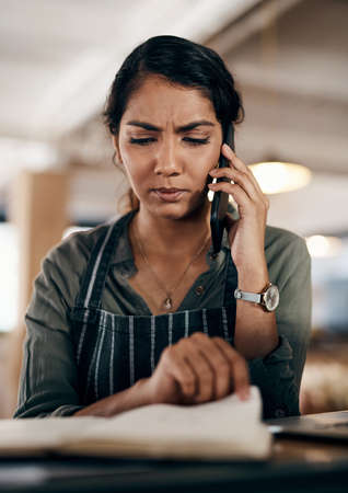 Young and confused business owner on the phone making a stock order for her cafe, restaurant or coffee shop. A female employee, manager or store owner looking at a book of products or inventory.の写真素材