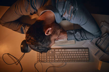 Hes hit shutdown during the late shift. High angle shot of a young businessman sleeping at his desk while working late in an office.の写真素材