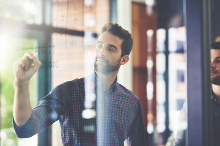 Theyve got a foolproof plan of action. two young businessmen brainstorming on a glass wall in an...
