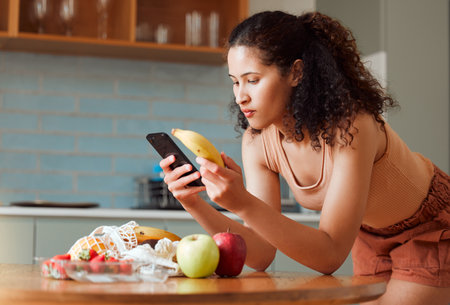 Woman reading phone, researching a diet with fresh fruit while relaxing in a kitchen at home. Young female searching for a recipe, cleanse or detox online. Lady checking nutritional value of a bananaの写真素材