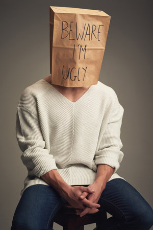 They think Im ugly but Im not. an unrecognizable man wearing a paper bag saying beware Im ugly while being seated against a grey background.の写真素材