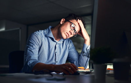 Tired, stressed businessman suffering from headache, working late night in the office. Serious, frustrated and overworked corporate male with headache staring at his screen in the dark.の写真素材