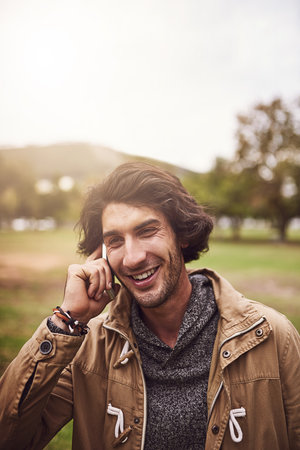 We can talk for hours. a cheerful young man talking on his phone while standing outside in a park.の写真素材