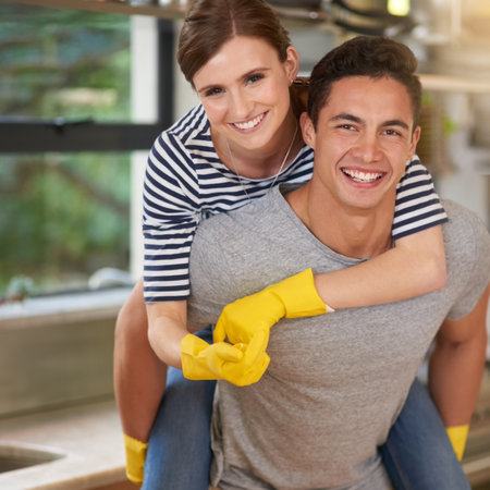 Who said spring cleaning had to be a chore. Portrait of happy young man giving his wife a piggyback ride while they clean their kitchen.の写真素材