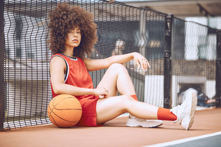 Relaxed, attitude and cool female basketball player relaxing outdoors on a court. Portrait of a trendy and fashionable black woman athlete sitting on the floor with a ball after trainingの写真素材