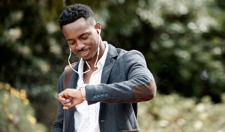 Checking the time on his watch, commuting and listening to music on earphones outside with a business man walking to his job or work. Feeling happy, positive and smiling while traveling outdoorsの写真素材