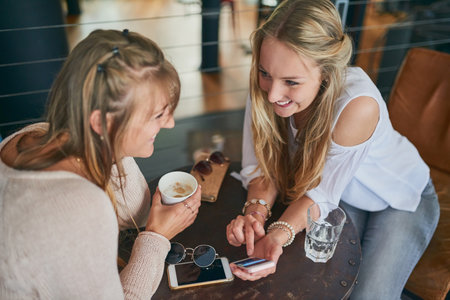 Catching up over coffee. High angle shot of two attractive young girlfriends looking at a cellphone while sitting in a cafe.の写真素材