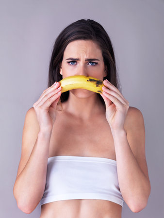 Shes one sad monkey...Studio portrait of an attractive young woman holding a banana against a purple background.の写真素材