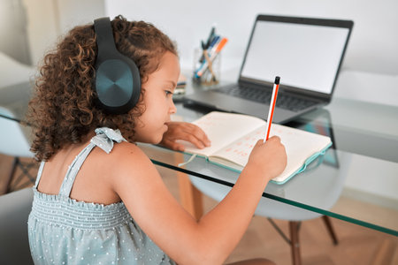 Homeschooling by a smart and clever little girl attending online or virtual class using a laptop and headphones. A young child writing and working or doing homework at home listening to musicの写真素材