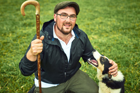 We are going to herd some sheep today. Portrait of a cheerful young farmer and his dog relaxing on a green field on a farm during the day.の写真素材