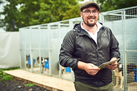 We breed the best dogs for farming here. Portrait of a cheerful young farmer standing with a digital tablet while a group of dogs can be seen relaxing in cages in the background.の写真素材