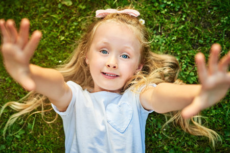Pick me up. Portrait of a cheerful little girl lying on grass stretching her arms out while looking at the camera outside.の写真素材